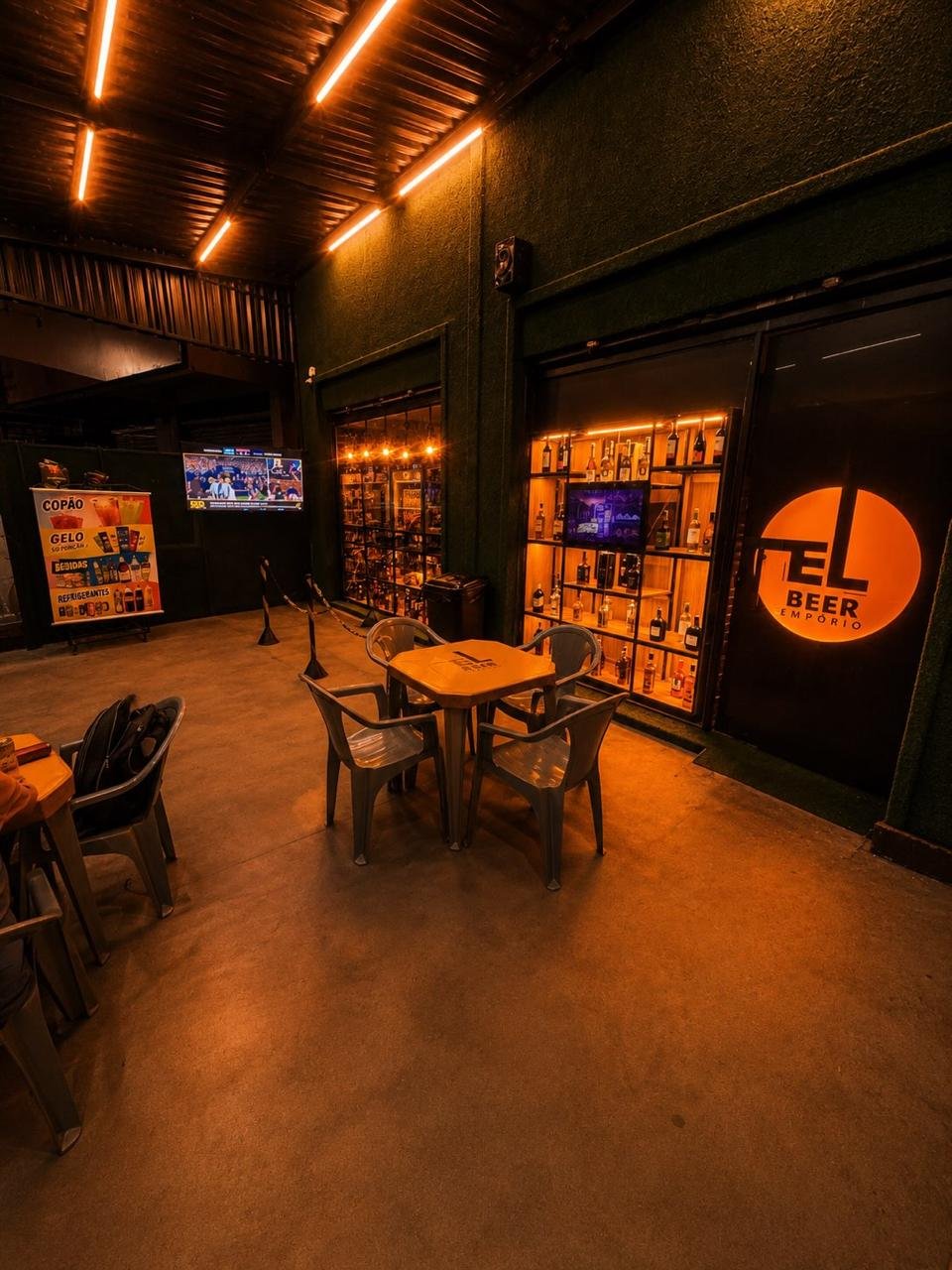 Espaço coberto do El Beer com mesas, TV ligada e a prateleira de destilados iluminada